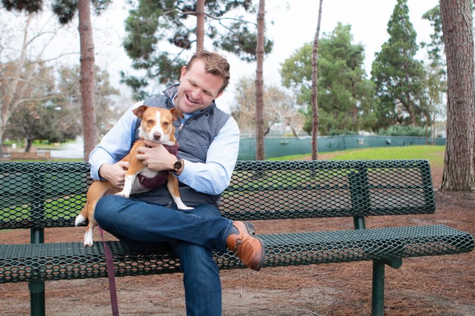 Steve Smith, candidate for Costa Mesa City Council District 5, smiles while holding a small tan and white dog on a park bench.